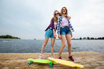 Two pretty smiling blond girls wearing checkered shirts and denim shorts are standing on the pier and having fun with the river in the background. Two bright longboards are in front of them. Sport and