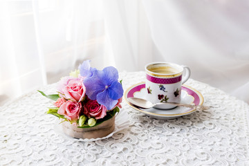 Beautiful soft pink roses and a blue hydrangea in the form of a capcake and a cup of coffee on a table covered with a white cloth. morning light through a white curtain