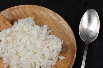 Boiled rice on a round wooden plate. Next to the plate lies a spoon. View from above. Dark background. Close-up.