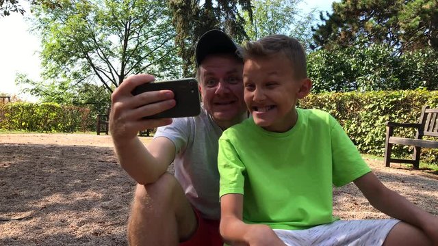 Father and son taking selfie and mug for phone camera in park at summer day. Good generations relationship: man and boy having fun. Dad and tween teenager act the ape with mobile