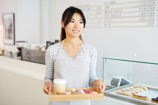 Young Asian waitress in workwear carrying fresh glazed eclairs and glass of cocktail on wooden tray