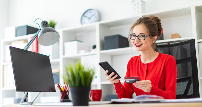 A young girl is sitting at the desk in the office, holding a bank card and phone in her hand.