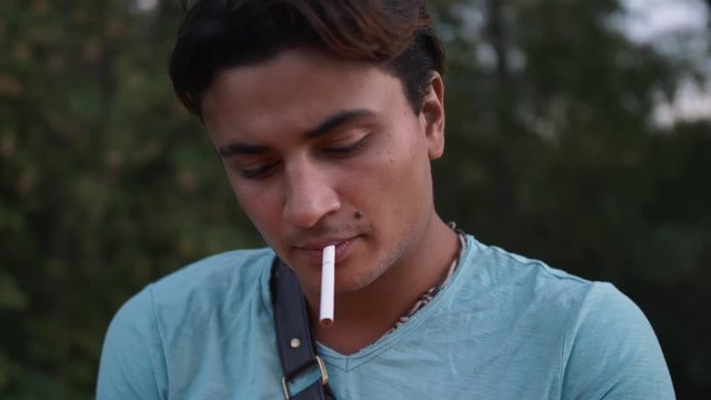 Handsome hispanic man takes a cigarette and light it up. Tired guy smokes a cigarette wearing blue shirt. Man has beauty spot birthmark right over lip