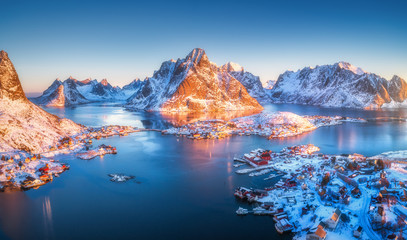 Aerial view of Reine at sunrise in winter. Top view of Lofoten islands, Norway. Panoramic landscape with blue sea, snowy mountains, high rocks, village with buildings, rorbu, sky reflected in water - obrazy, fototapety, plakaty