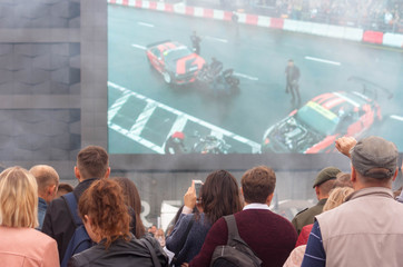 A view of a number of spectators who watch the motor show on the background of a large screen that broadcasts the spectacle in real time.