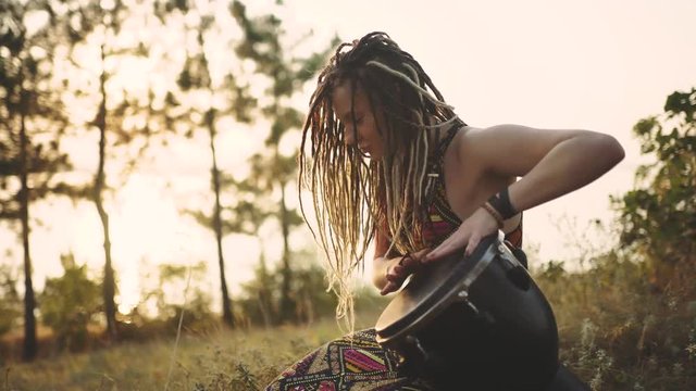 Beautiful young hippie woman with dreadlocks playing on djembe. Funky woman drumming in nature on an ethnic drum at sunset or sunrise