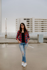 Obraz premium Cute Dark Haired Female Model Posing in Ripped Jeans and Plaid Shirt on Top Floor of Parking Garage