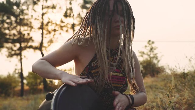 Beautiful young hippie woman with dreadlocks playing on djembe. Funky woman drumming in nature on an ethnic drum at sunset or sunrise