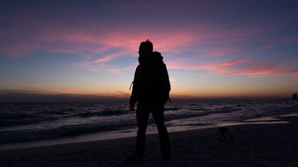 Guy stands in front of dramatic sunset over the ocean on a beach covered with waves.