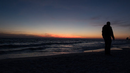 Guy stands in front of dramatic sunset over the ocean on a beach covered with waves.