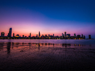 Gorgeous long exposure Chicago skyline photograph just after sunset with pink, purple and blue colors in the sky and reflecting on the water and wet pavement as the highrise building lights turn on.