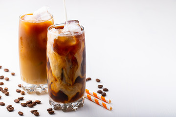 Ice coffee in a tall glass with cream poured over and coffee beans. Cold summer drink on white background with copy space