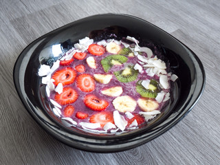 Vibrant colorful and fresh fruit topped acai bowl or smoothie bowl with purple colored blueberry smoothie with strawberries, banana, kiwi and coconut flakes lining a black bowl on woodgrain background