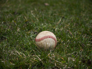 Close up sports background image of an old used weathered leather baseball ball laying in the grass field outside showing intricate detailing and red laces.