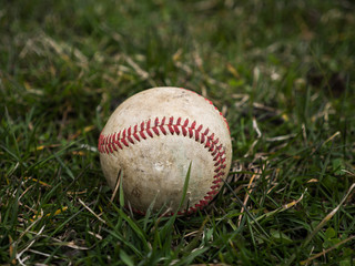 Close up sports background image of an old used weathered leather baseball ball laying in the grass field outside showing intricate detailing and red laces.