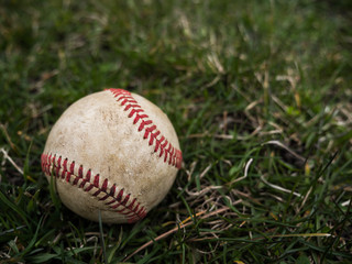 Close up sports background image of an old used weathered leather baseball ball laying in the grass field outside showing intricate detailing and red laces.
