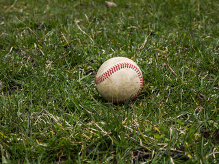 Close up sports background image of an old used weathered leather baseball ball laying in the grass field outside showing intricate detailing and red laces.