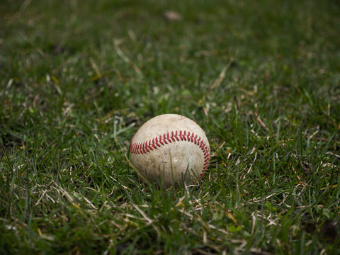 Close up sports background image of an old used weathered leather baseball ball laying in the grass field outside showing intricate detailing and red laces.