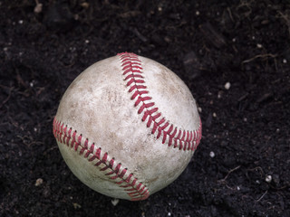 Close up sports background image of an old used weathered leather baseball laying in a pile of brown dirt showing intricate detailing and red laces.