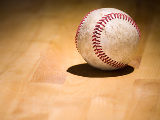 Close up sports background image of an old used weathered leather baseball showing intricate detailing and red laces sitting on a wood butcher block counter with lighting from behind and dark shadows.