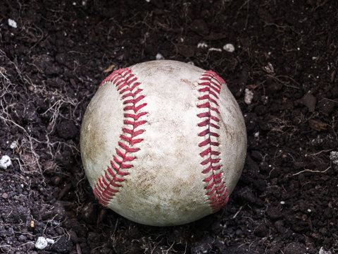 Close up sports background image of an old used weathered leather baseball laying in a pile of brown dirt showing intricate detailing and red laces.