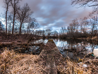 View looking down a wood plank to a weathered pier sticking out into the calm water of a pond at sunset with pink and blue fluffy clouds above and trees reflecting in the water.