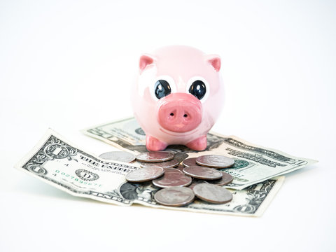 Photograph of a cute pink ceramic piggy bank sitting on top of a pile of united states dollar bills and coins isolated on a white background.