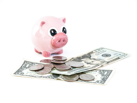 Photograph of a cute pink ceramic piggy bank sitting next to a pile of united states dollar bills and coins isolated on a white background.