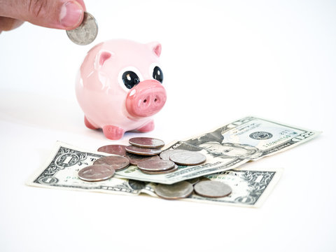 Photograph of a Caucasian male hand holding a United States quarter coin inserting it in a pink ceramic piggy bank sitting on dollar bills and coins isolated on a white background.
