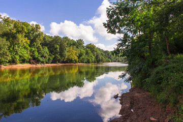 Reflections of clouds on river lined with green trees © Adam