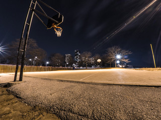 Wide angle photograph of a concrete exterior basketball court with hoops at either end, street lights with residential highrise apartments in the distance in the dark night sky in Chicago. © Joseph Kirsch