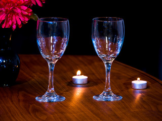 Romantic background photograph of two empty wine glasses displayed on a dark wood grain table with tea candles and red flowers in the background.