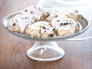 Food photograph of several freshly made homemade blueberry scones with white lemon and powder sugar glaze sitting on a glass cake stand on a wood table for a beautiful and delicious background image.