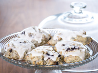 Food photograph of several freshly made homemade blueberry scones with white lemon and powder sugar glaze sitting on a glass cake stand on a wood table for a beautiful and delicious background image.