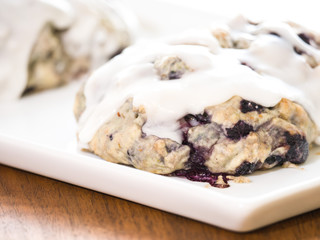 Close up of homemade blueberry scone pastries fresh out of the oven with melted white lemon and powder sugar glaze sitting on a white serving tray on a wood grain table.