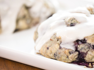 Close up of homemade blueberry scone pastries fresh out of the oven with melted white lemon and powder sugar glaze sitting on a white serving tray on a wood grain table.