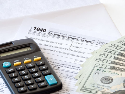 Close-up photograph of a hard copy of an individual income tax return with black calculator and 20 dollar bills piled up representative of United States federal tax cuts and savings this year.