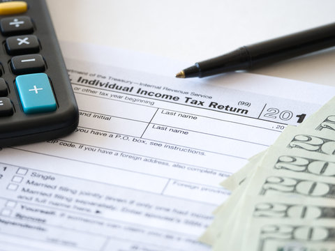 Close-up photograph of a hard copy of an individual income tax return with black calculator and 20 dollar bills piled up representative of United States federal tax cuts and savings this year.