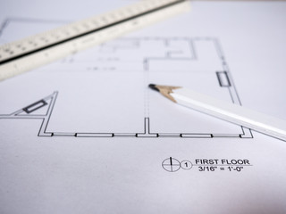 Photograph of generic architectural residential floor plan drawings printed on white paper set on top of a wood desk in a architect studio with scale ruler and carpenters pencil.