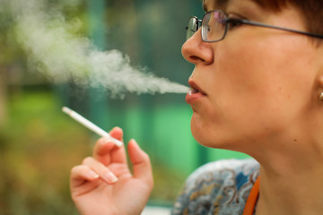Fototapeta premium woman smokes thin cigarette, exhaling smoke. Female hand with smoking cigarette on blurred green park background