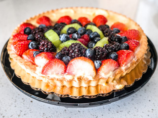 Closeup photograph of a beautiful and colorful vibrant fruit tart dessert with blueberry, strawberry, kiwi and blackberry fruit topping and browned fluted crust edge set on a kitchen counter top.