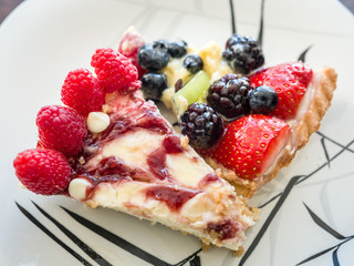Closeup photograph of a moist piece of white chocolate raspberry cheesecake and mouth watering fruit tart on a white and black dessert plate with vibrant colors.