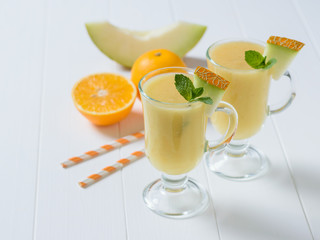 Two glass mugs with melon smoothie, orange and melon on a white wooden table.
