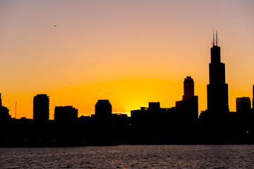 Naklejka na ściany i meble Chicago skyline picture during beautiful orange yellow sun as it lowers below the building silhouettes and the water of lake Michigan in the foreground