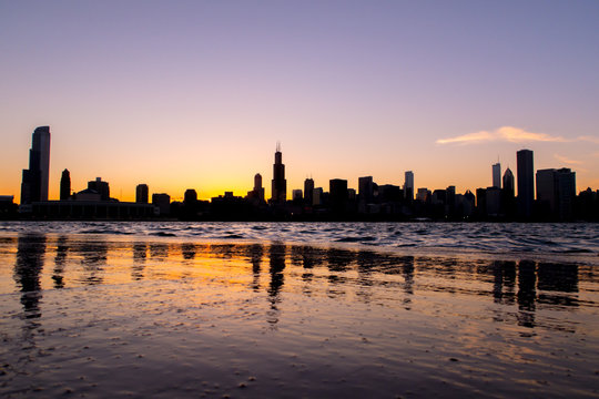 Chicago skyline picture during beautiful orange yellow sun as it lowers below the building silhouettes and reflections in the water of lake Michigan in the foreground
