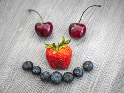 Silly smiley face composed with ripe fruit including cherry eyes, strawberry nose, and blueberry mouth on a gray wood textured background