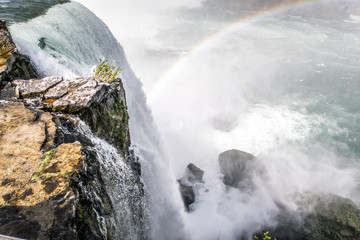 Beautiful view looking over the edge of Niagara Falls waterfall with rainbow forming in the mist below as water hits the boulders below with rocky cliff edge in foreground