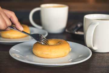 A hand cutting a piece of donut by fork in a white plate with coffee cups on table