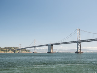 A beautiful view of the expensive and famous San Francisco Oakland Bay Bridge with suspension cables and towers protruding from the San Francisco Bay with water below and clear blue sunny sky above.