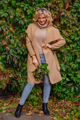 Young beautiful woman in sweater and coat stands next to the background of wild grapes holding bouquet of autumn leaves